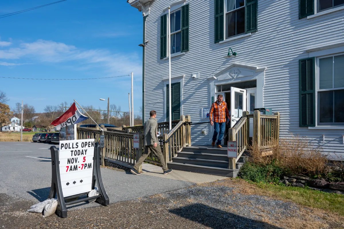 A person wearing a plaid shirt exits a polling station. Another person approaches the building. A sign reads, "Polls Open Today Nov. 5, 7AM - 7PM.