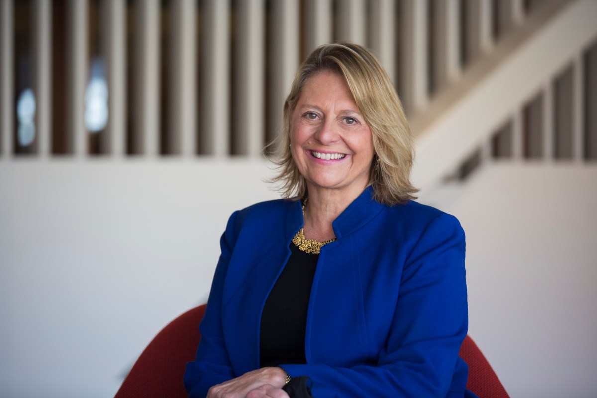 A woman with blonde hair wearing a blue blazer and black top sits on a red chair, smiling at the camera. There is a staircase with white vertical railings in the background.