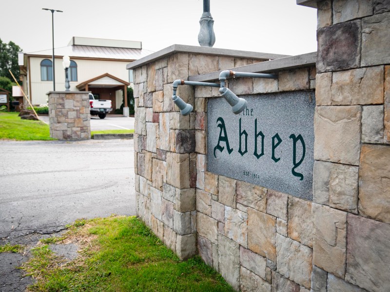 Stone sign with "The Abbey" engraved, mounted on a stone wall at the entrance to a property with buildings in the background.