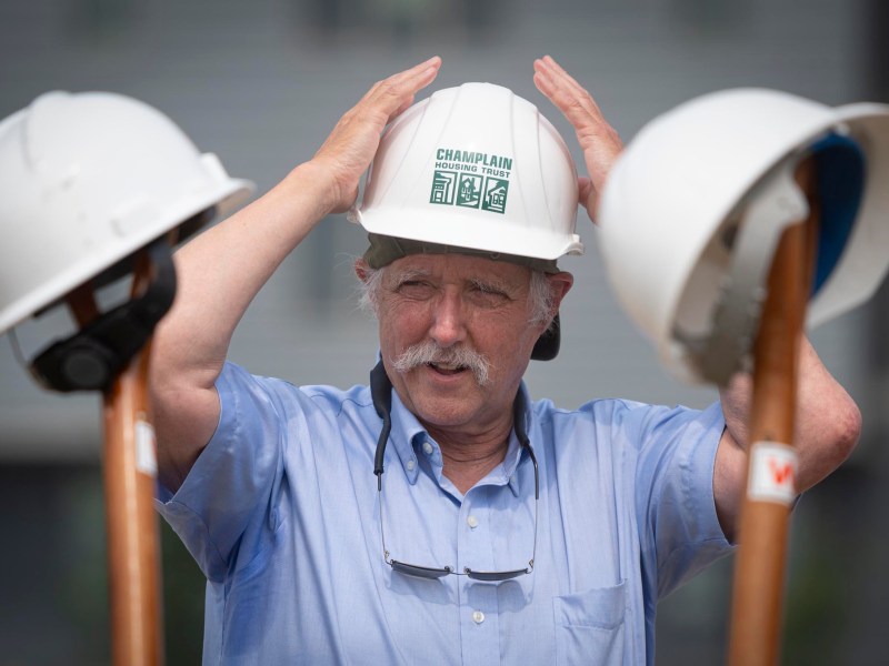 An older man in a blue shirt adjusts a hard hat labeled "CHAMPLAIN," with two other hard hats resting on shovels in the foreground.