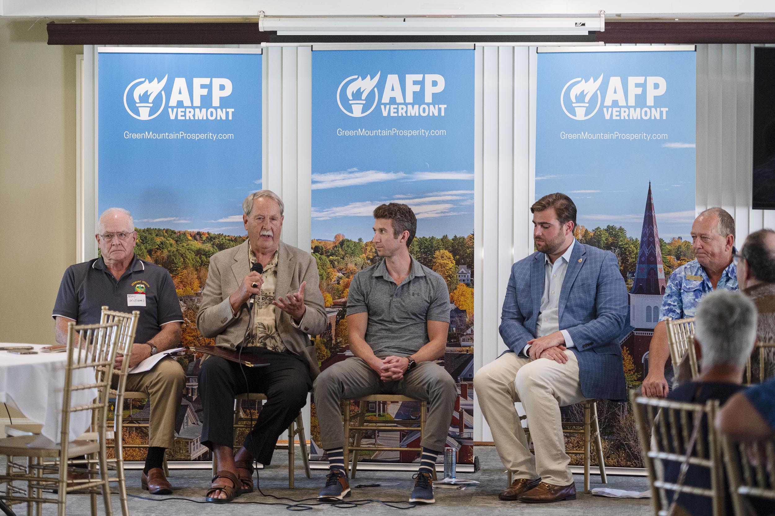 Five men are seated on a panel in front of AFP Vermont banners, with one man speaking into a microphone during an event.