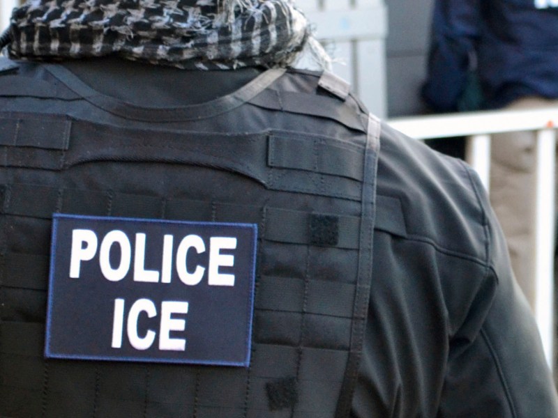 A close-up of a person wearing a vest labeled "Police ICE," standing outdoors near a metal barrier.