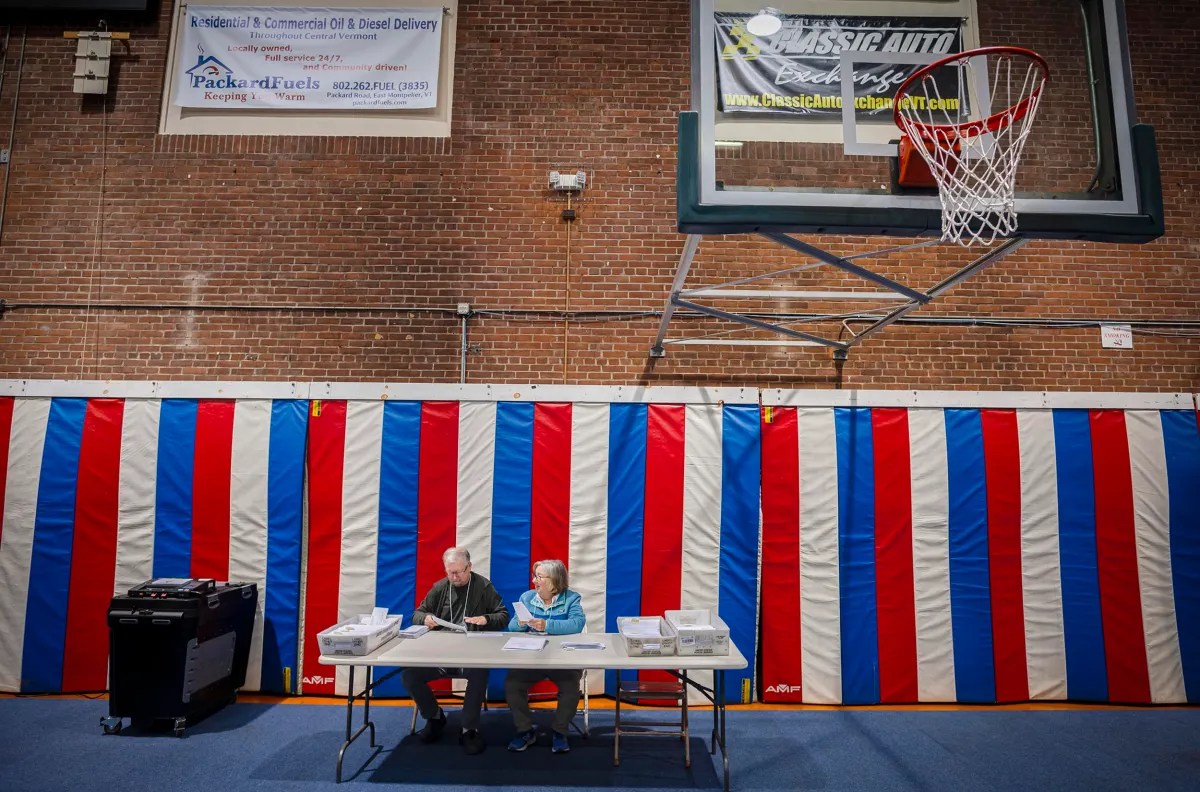 Two people sit at a table in a gymnasium near a basketball hoop, with papers and a voting machine nearby. Red, white, and blue striped curtains hang in the background.