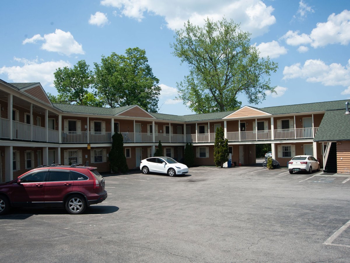 A parking lot with three cars in front of a two-story motel. The building has exterior staircases and balconies. Trees and clouds are in the background.