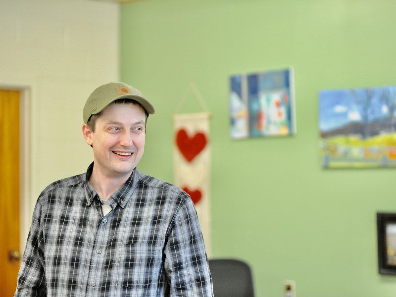 A man in a plaid shirt and cap stands indoors, smiling, with paintings and a heart-themed wall hanging in the background.