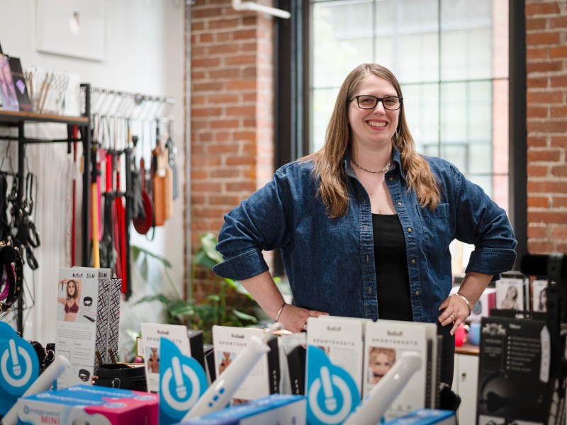 A woman stands smiling behind a display of adult products in a store with brick walls, shelves, and large windows in the background.