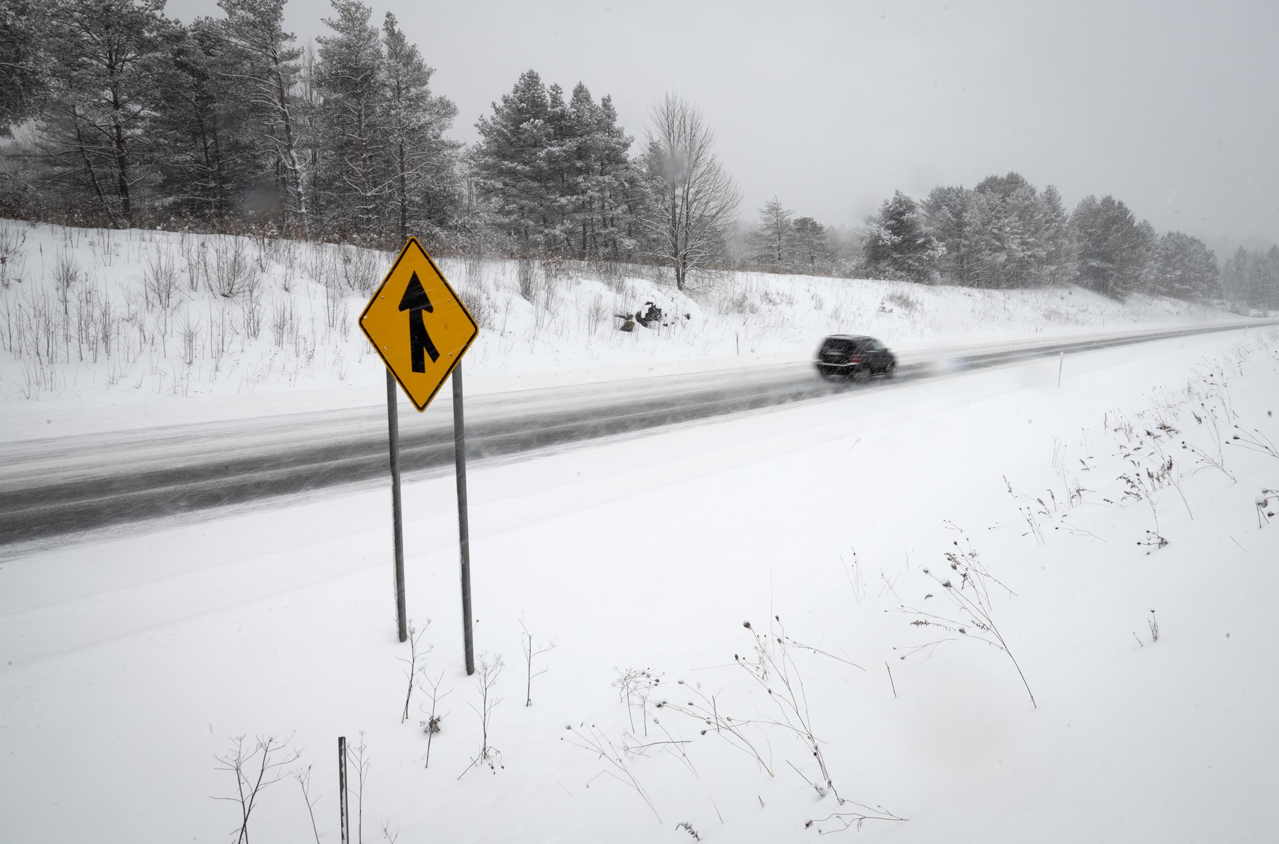 Car drives on a snow-covered road past a yellow merge sign. Snow blankets the landscape and trees, creating a wintry scene.