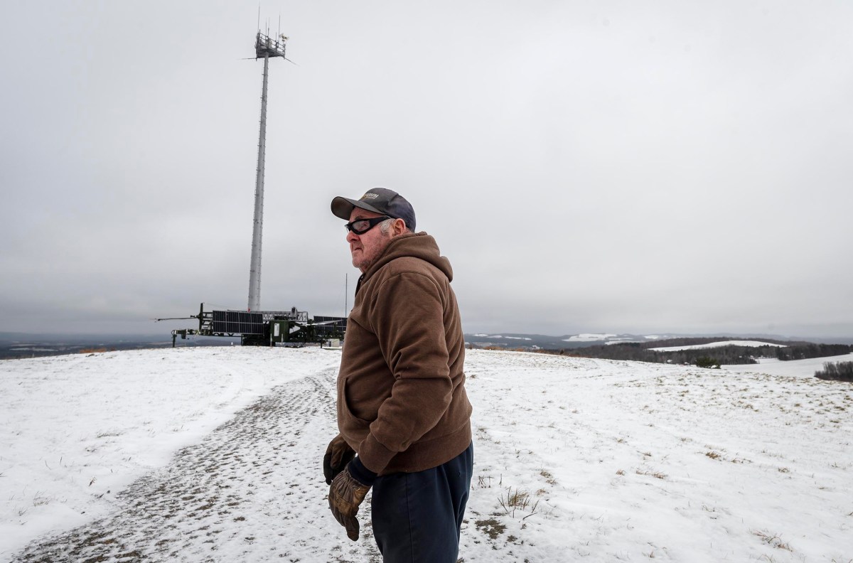 A man in a brown jacket and cap stands on a snowy hill near a tall communication tower under a cloudy sky.