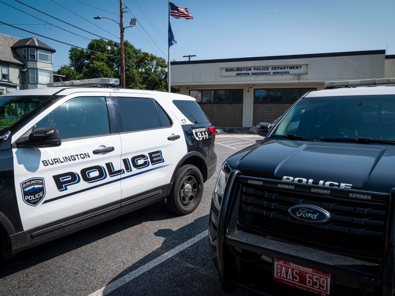 Two police SUVs are parked in front of the Burlington Police Department building, which has American and state flags flying above it.