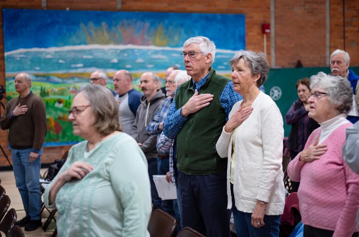 A group of people stand with hands on their chests in a room with chairs, facing a large landscape mural on the wall.