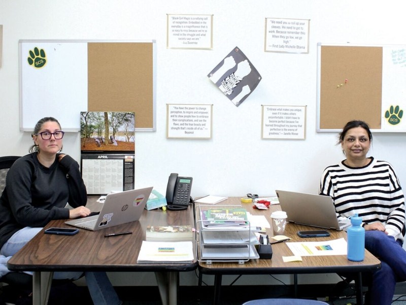 Two women sit at separate desks in an office with bulletin boards, wall quotes, a calendar, and various office supplies visible on the tables.