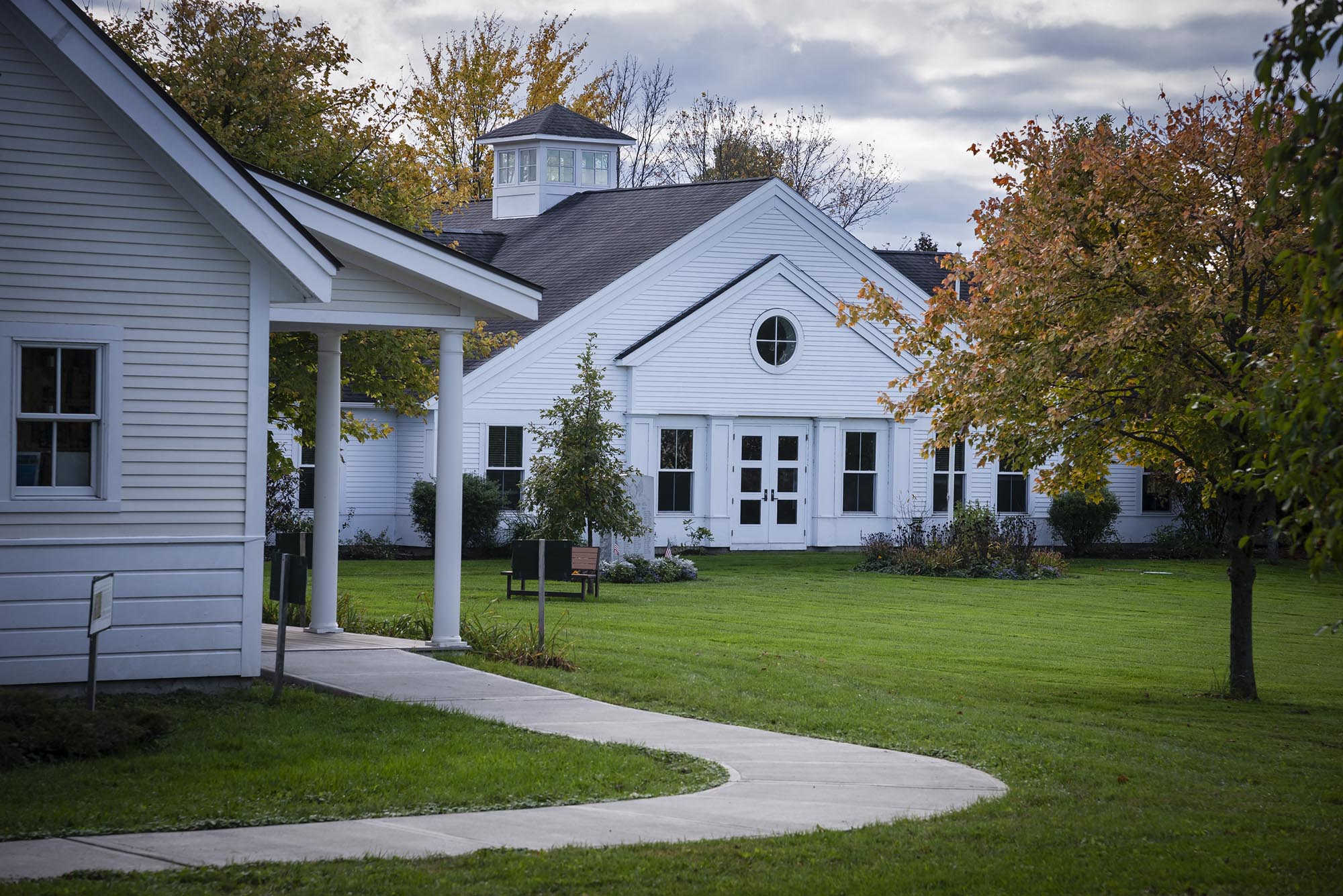 A sidewalk winds through white buildings, fall foliage and a green lawn.