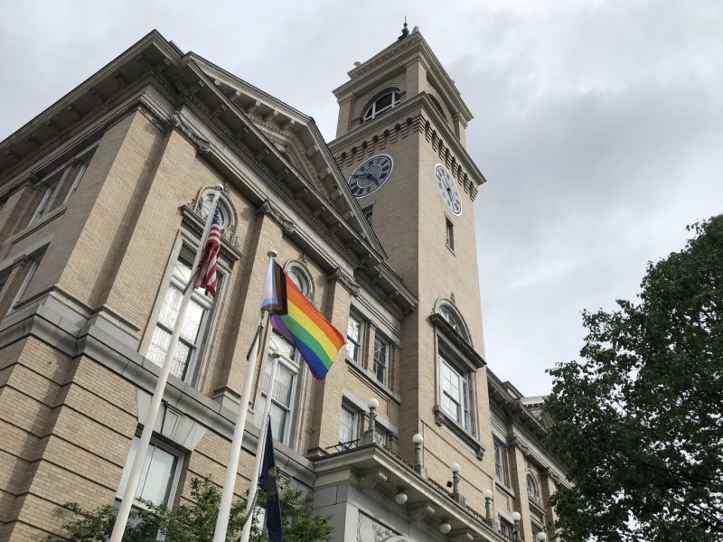 A historic building with a clock tower features two flagpoles. One flag is an American flag and the other is a rainbow pride flag. A tree is visible to the right.