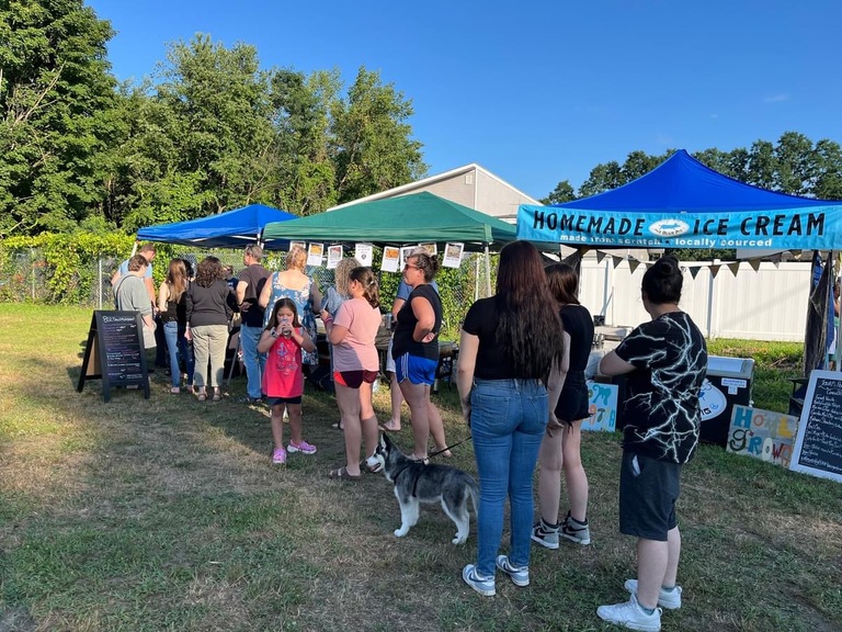 People are standing in line at a homemade ice cream stand with blue and green tents at an outdoor event on a sunny day. A dog is also present. Trees and a building are visible in the background.