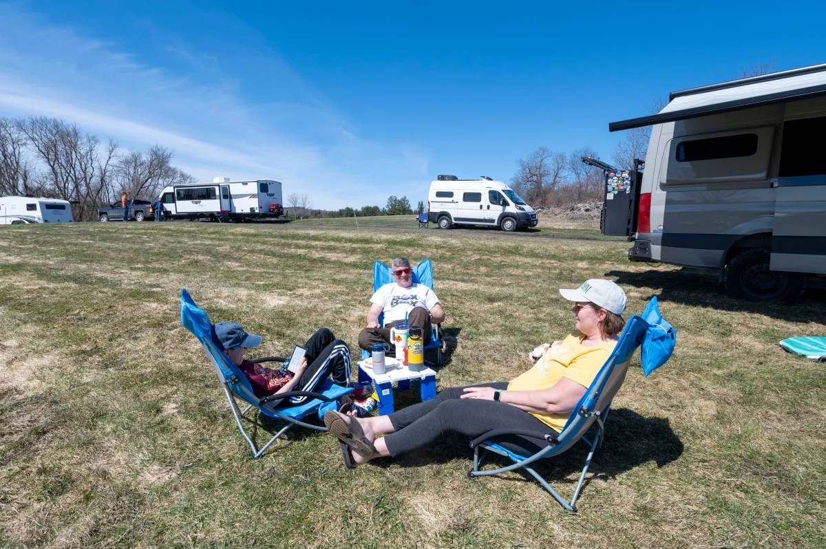 Two people relaxing in camping chairs in front of a recreational vehicle in a grassy area with other vehicles in the background.