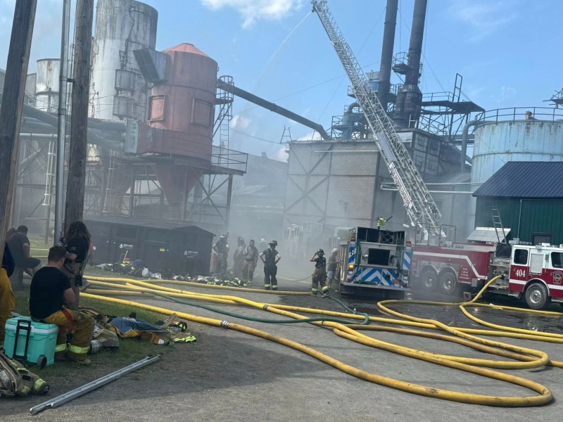 Firefighters respond to an industrial fire with hoses and ladder trucks as smoke rises from large factory structures in the background.
