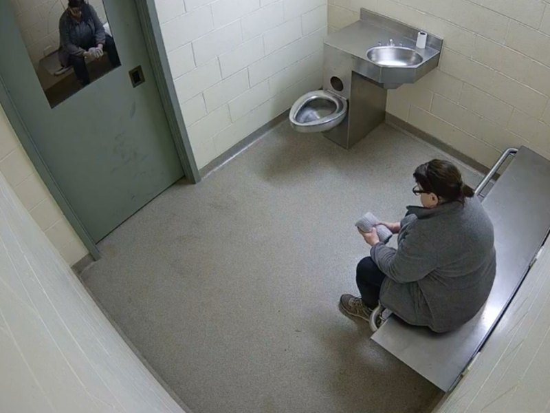 A woman sits alone on a metal bench in a sparse jail cell, holding papers, with a toilet and sink visible in the corner.