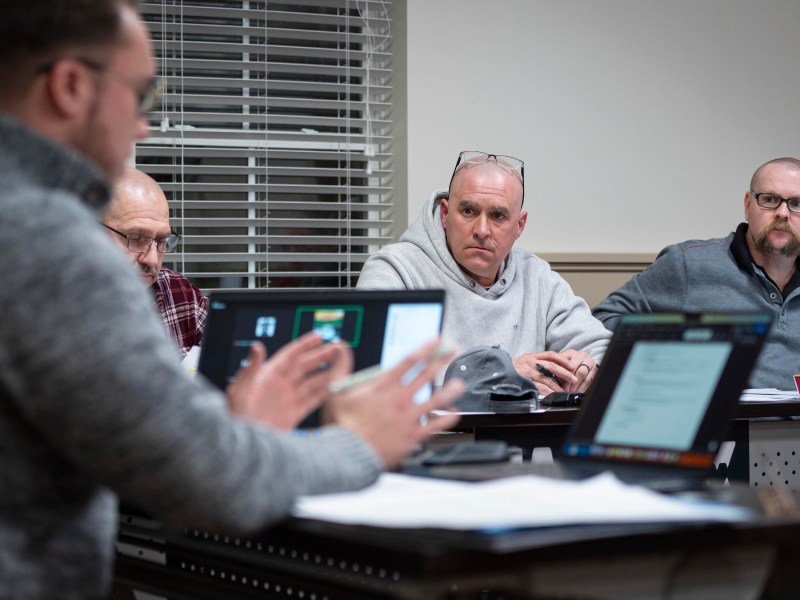Four men sit at a conference table engaging in discussion, with laptops open in front of them. One man is speaking while the others listen intently.