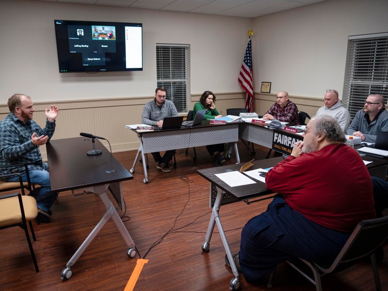 A man speaks at a table to a group of six people during a meeting. A screen shows a virtual participant. An American flag is in the background. Papers and laptops are on the tables.