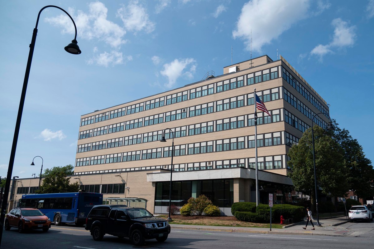 A large, rectangular office building with many windows, an American flag outside, cars and a blue bus parked nearby, and a partly cloudy sky overhead.
