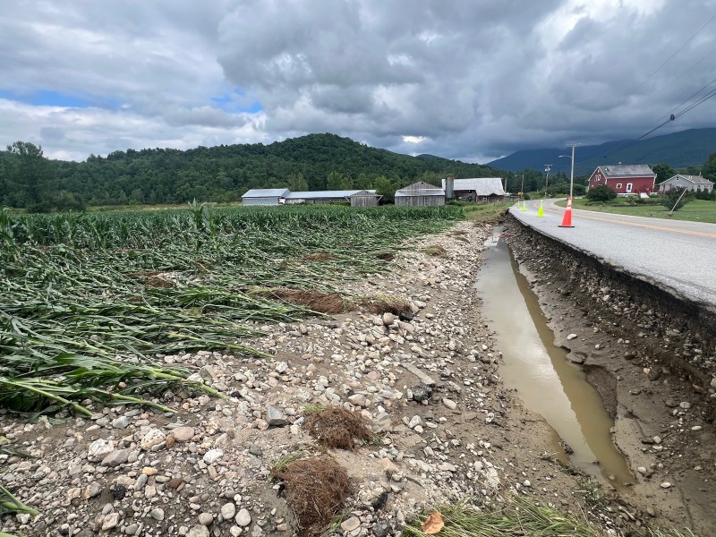 Roadside erosion with a partially collapsed road, adjacent to a flooded ditch and damaged crops, set against a backdrop of mountains and farm buildings under cloudy sky.