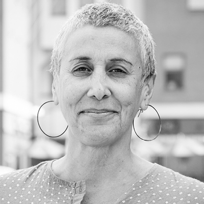 A person with short gray hair and hoop earrings smiles slightly while standing outdoors.