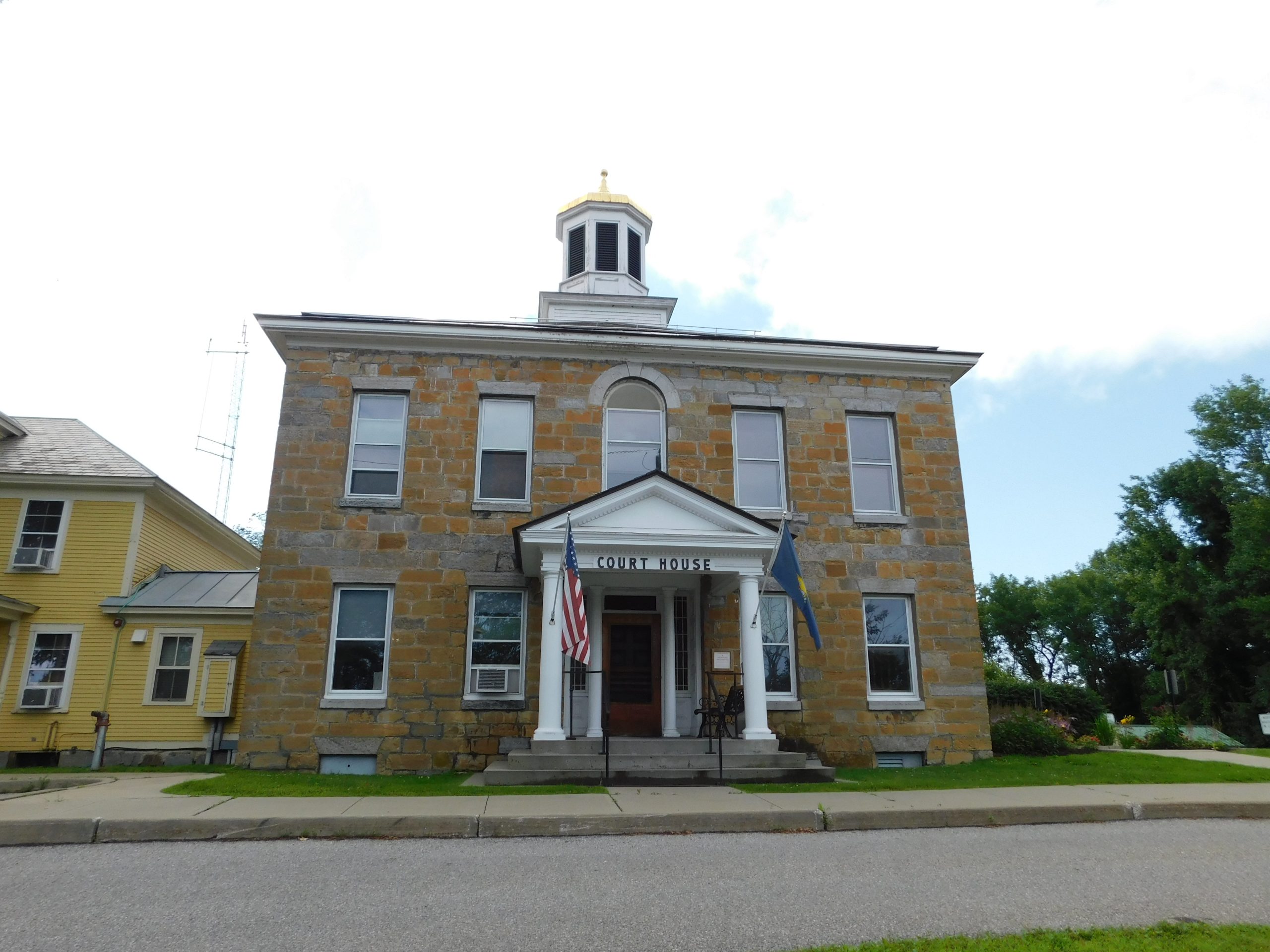A two-story stone courthouse with a small central tower, American flag, and another flag on its front. Steps and pillars lead to the entrance. Adjacent is a yellow house. Trees and a blue sky are in the background.