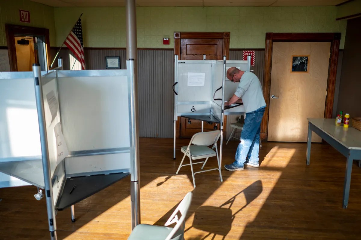A person votes in a polling booth in a room with wooden floors and chairs. An American flag is visible in the background.