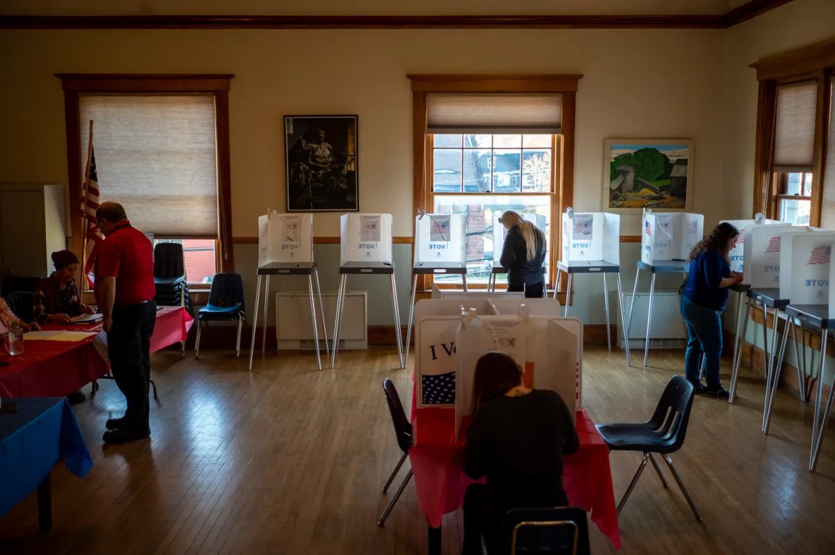 People voting in a polling station with voting booths along the walls and others seated at tables. Flags and artwork decorate the room.