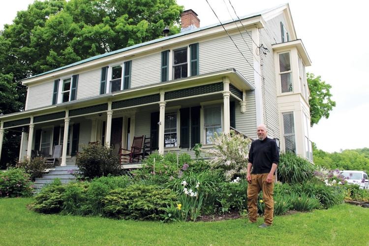 A man stands in front of a two-story white house with a large porch, surrounded by green plants and flowers on a cloudy day.