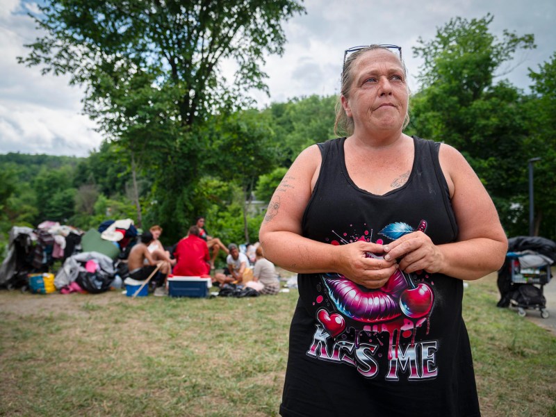 A woman stands outdoors in front of a group of people sitting near tents and bags, with trees and greenery in the background.
