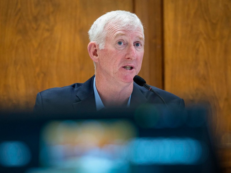 A man with gray hair speaks into a microphone. He is wearing a dark suit and is seated in front of a wooden background.