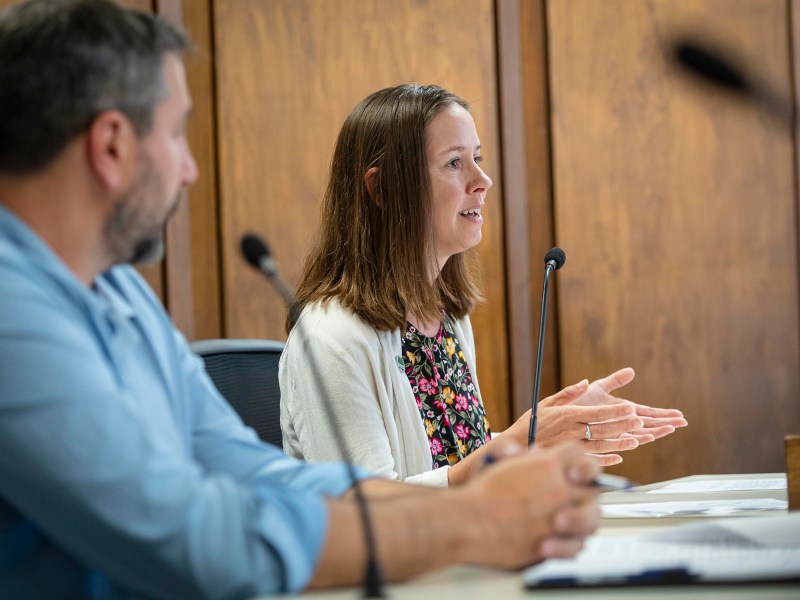 Two individuals are seated at a table, engaged in a discussion. The woman on the right is speaking, gesturing with her hands, while the man on the left listens attentively. They are in a wood-paneled room.