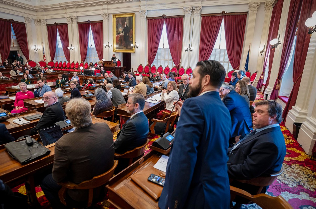 A legislative chamber with people sitting at desks, dressed in business attire, engaged in discussion. Red curtains and a large painting are visible in the background.
