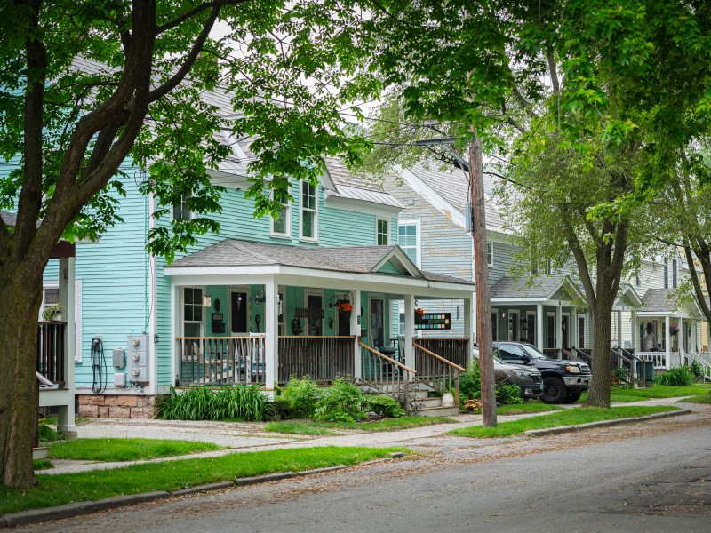 Row of pastel-colored houses with front porches, trees lining the street, and parked cars along the curb in a quiet suburban neighborhood.