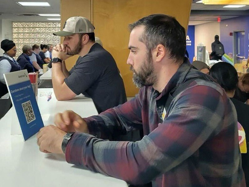 Two men sit at a white counter in an airport. One is looking at a sign with a QR code while others are seated in the background.