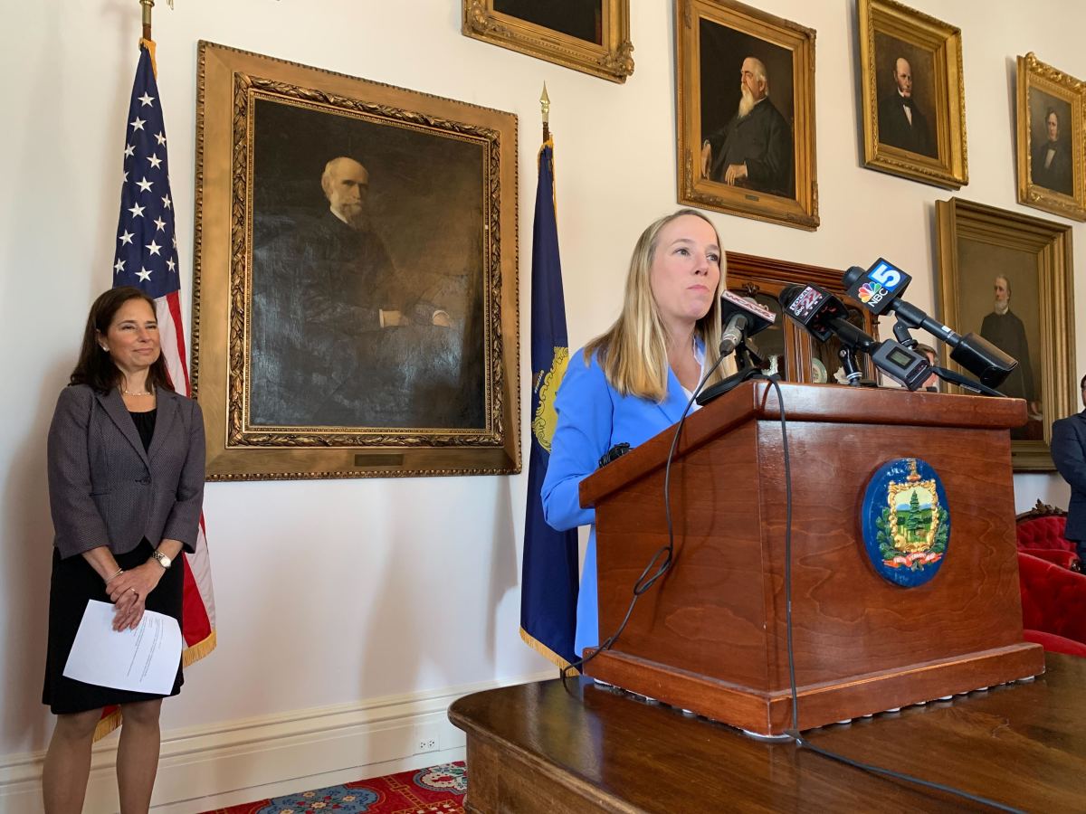A woman delivers a speech at a podium with microphones while another woman watches on, with portraits and an american flag in the background.