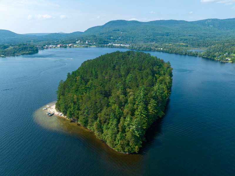 Aerial view of a densely forested island surrounded by a lake, with nearby shores in the background and distant hills under a blue sky.