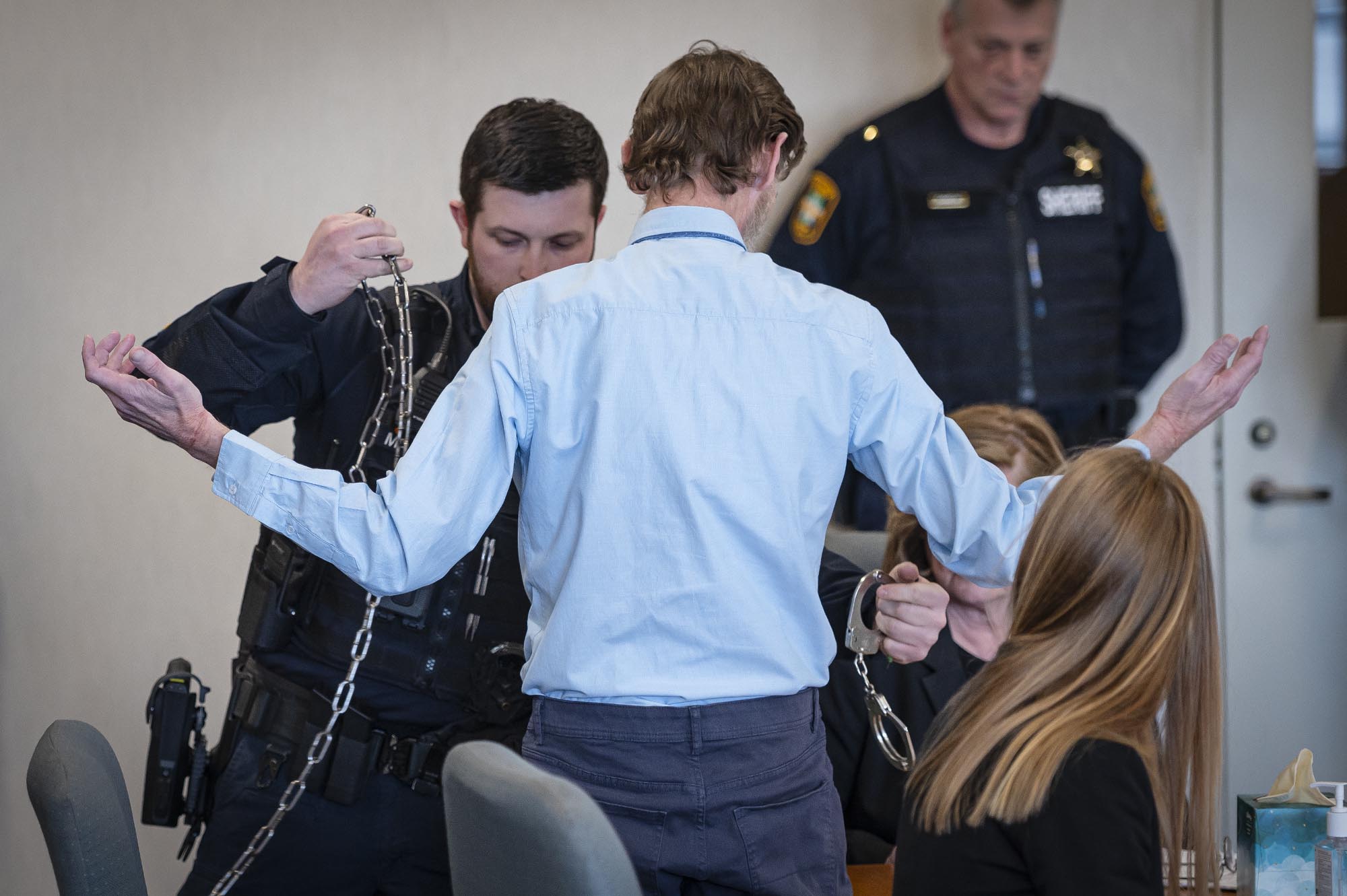 A police officer handcuffs a man in a blue shirt in a courtroom while others watch.