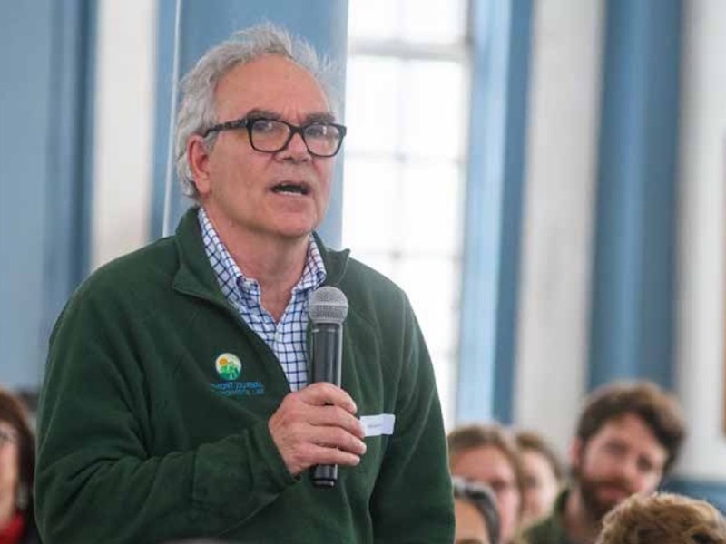 A man with glasses speaks into a microphone while standing in a room filled with seated people.
