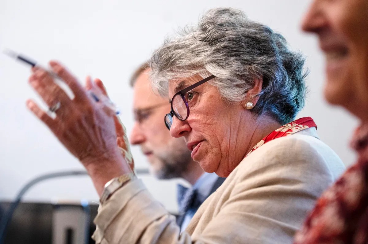 Older woman with gray hair and glasses speaks while gesturing with her hand; two other people are partially visible beside her.