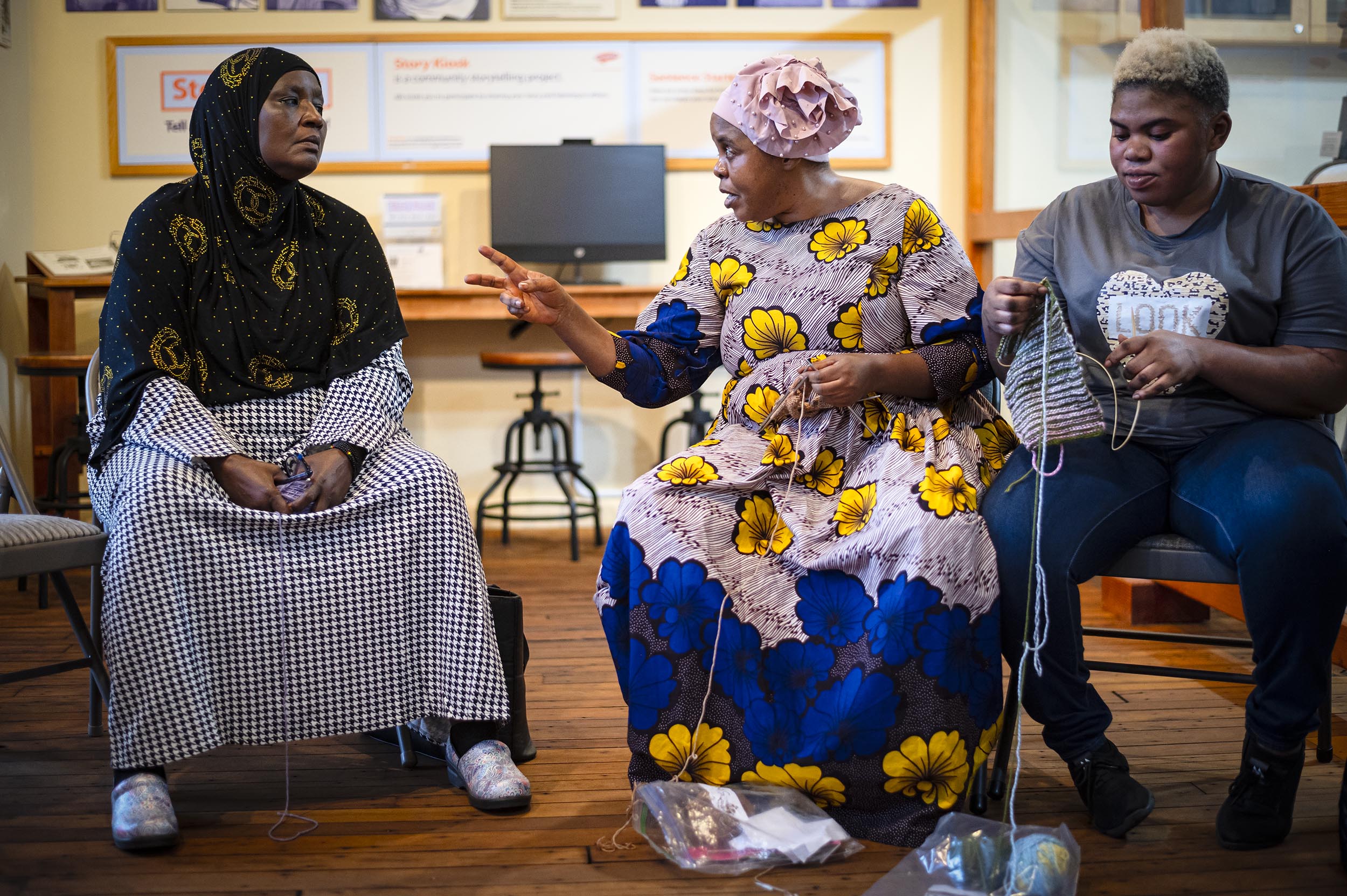 Three women sit together, knitting and talking. The woman in the middle, wearing a colorful dress, gestures with her hand while speaking. The other two women are focused on their knitting.