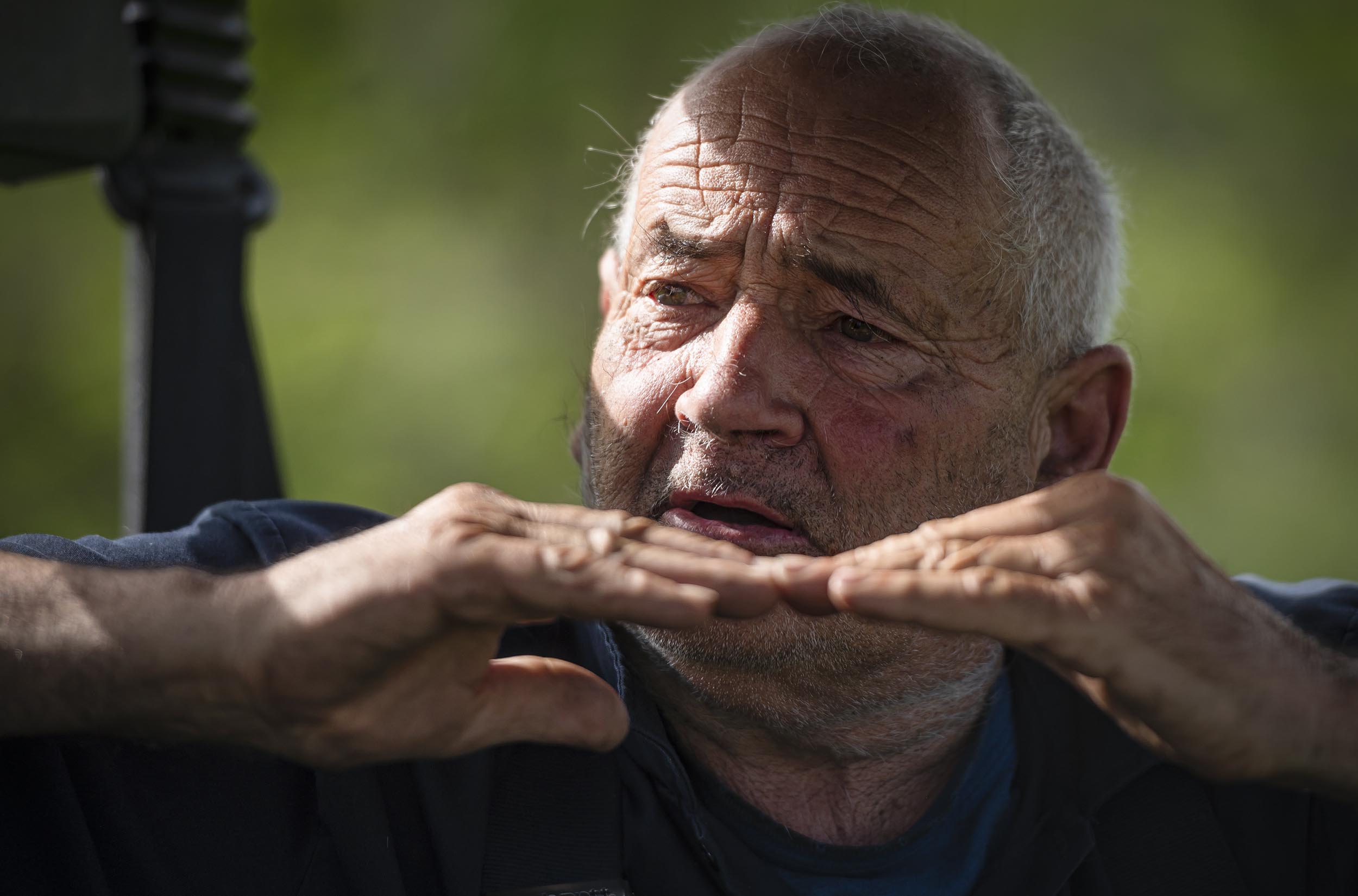 Close-up of an elderly man with short white hair and a rugged face, gesturing with hands at face level while outdoors. He wears a dark shirt, and the background is blurred with greenery.