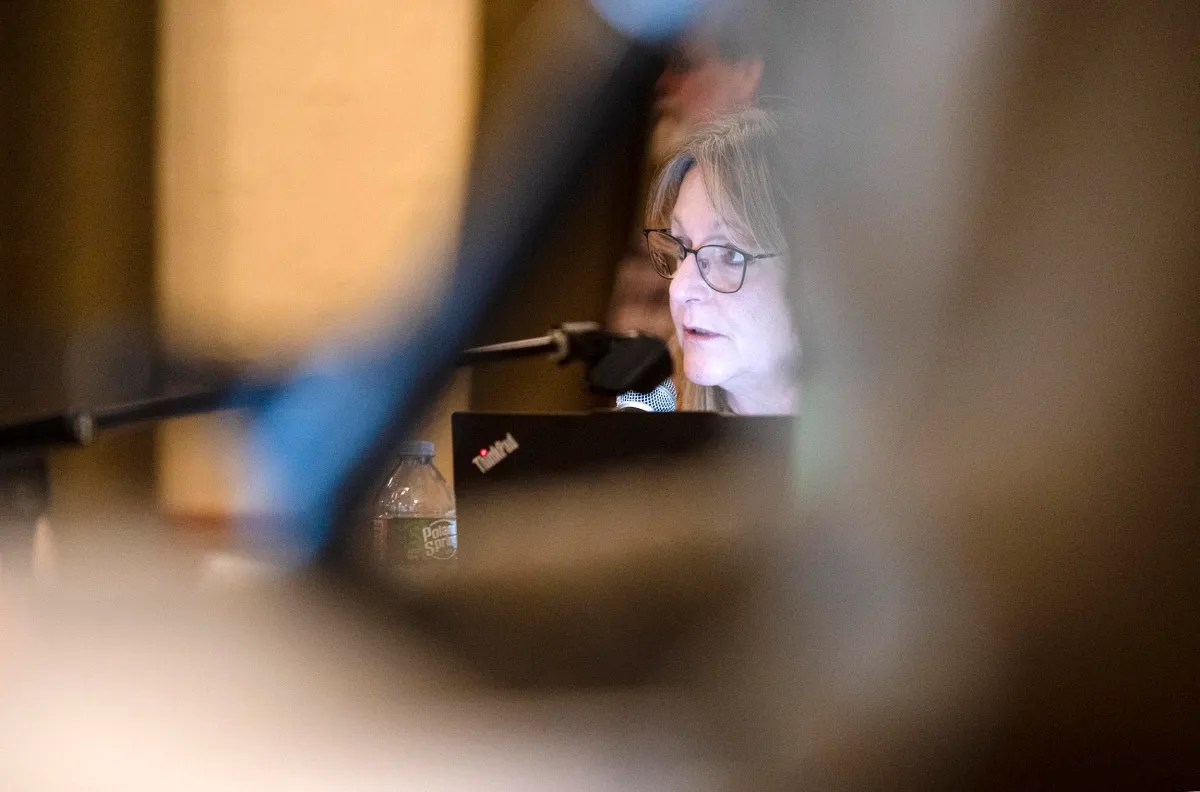 A woman speaking into a microphone while looking at a laptop, with a water bottle nearby, partially obscured by objects in the foreground.