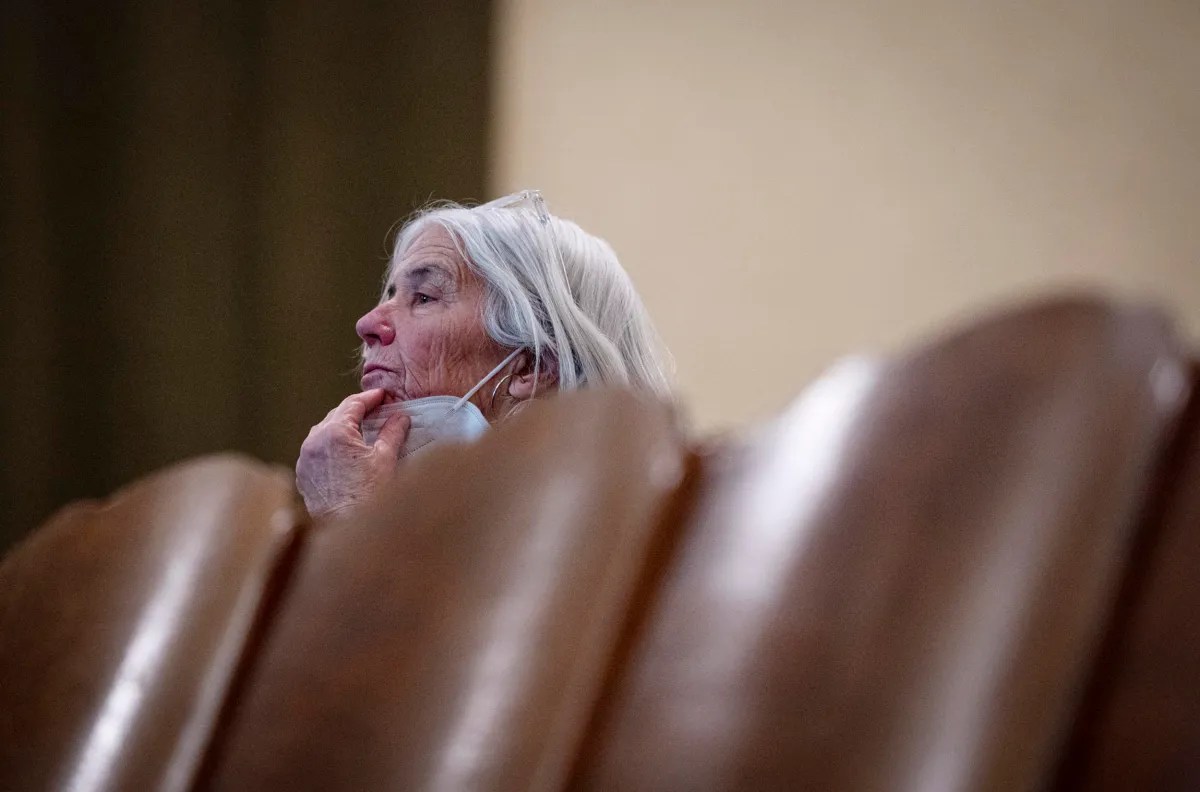 An elderly woman with white hair sits pensively in a row of brown seats, partially visible, wearing glasses and a face mask under her chin.