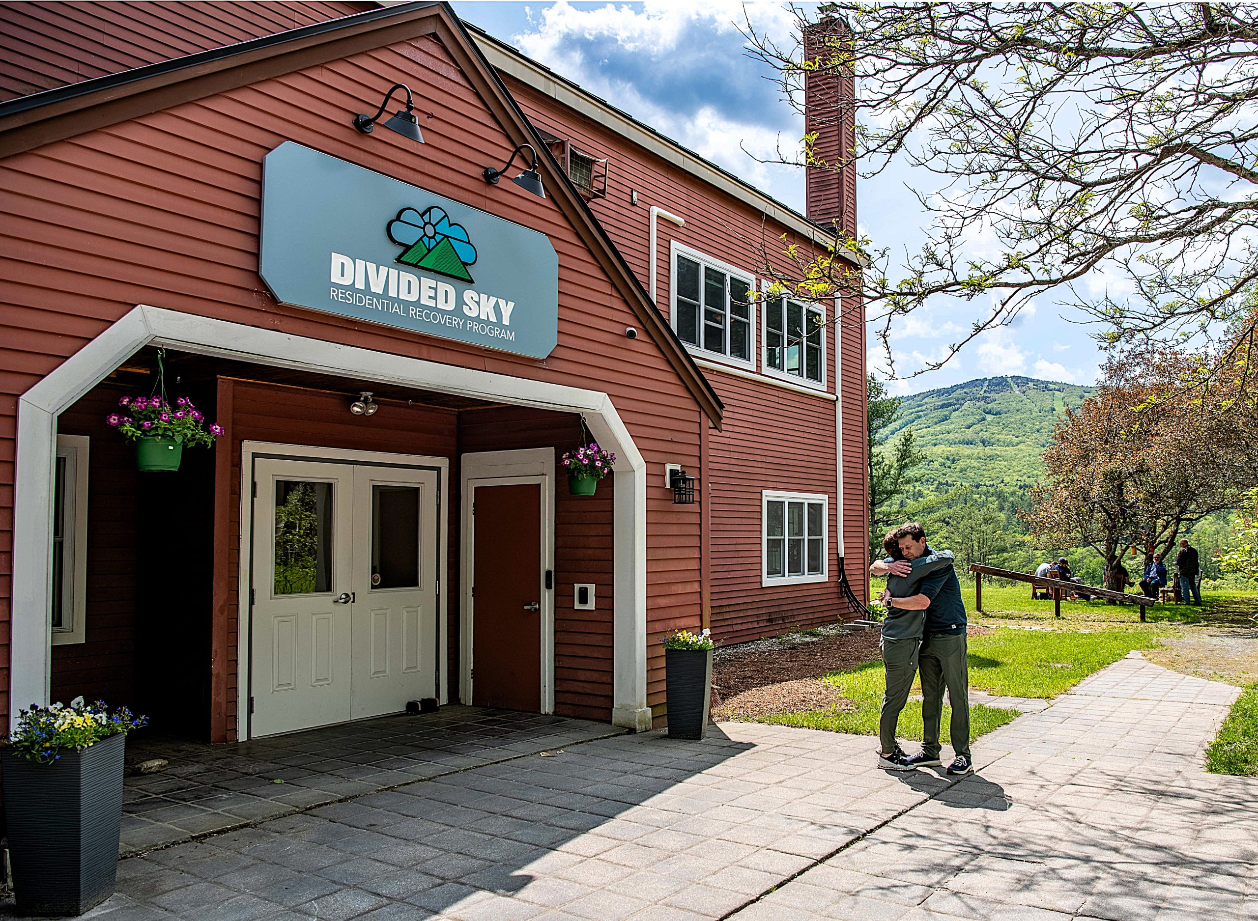 Two people hug outside the entrance of a red building labeled "Divided Sky Residential Recovery Program," with greenery and mountains visible in the background.