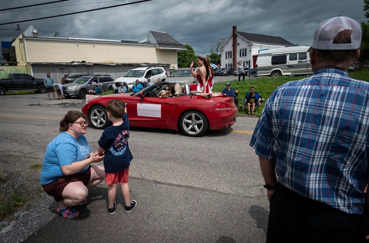 A red convertible with people standing inside passes by spectators, including a woman and child kneeling by the roadside and several people seated on the grass, during a cloudy day parade.
