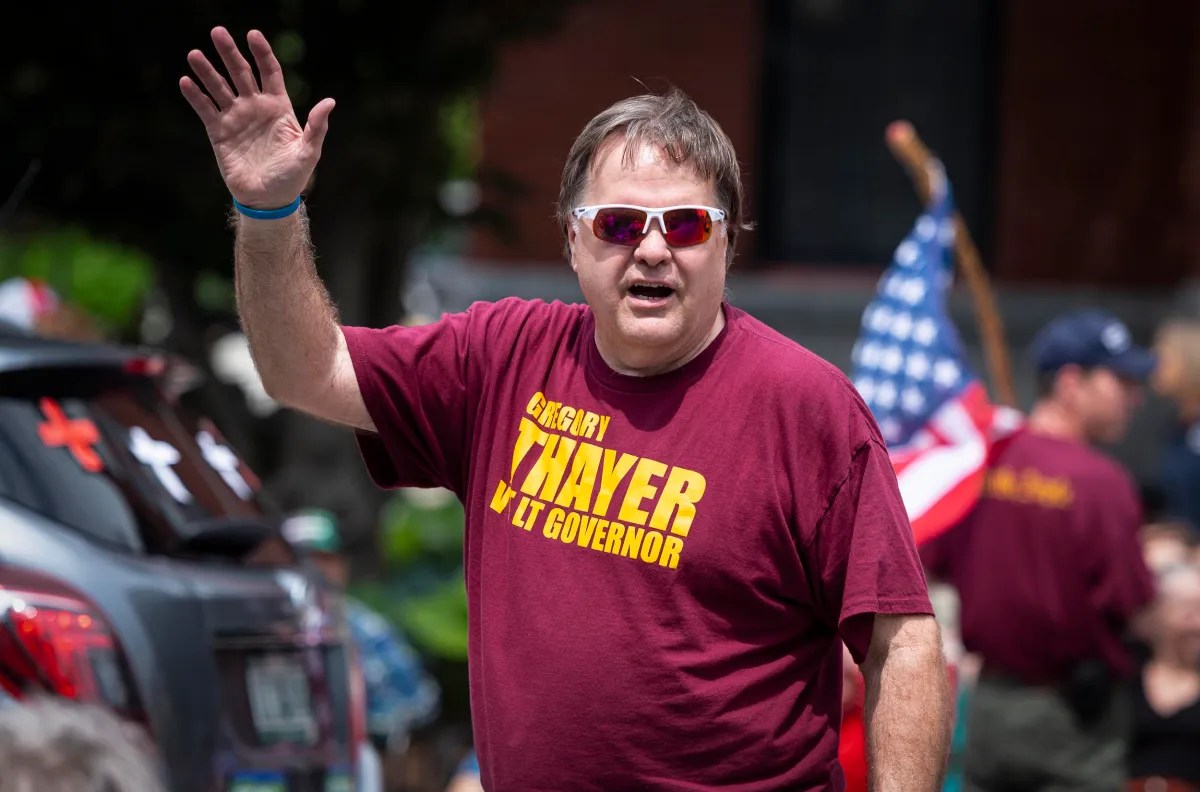 A man wearing a maroon "Gregory Thayer for Lt Governor" shirt waves his hand. He is outdoors, and an American flag is visible in the background.