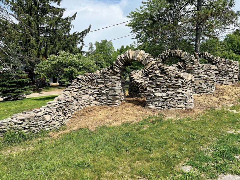 A dry stone wall forms a series of arches in a grassy outdoor area with trees and power lines in the background.