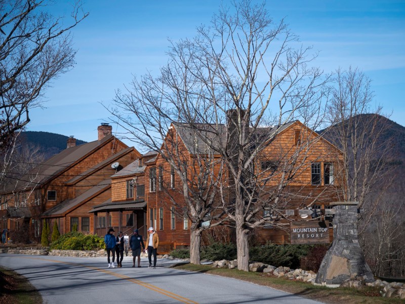 A group of people walk along a road beside rustic buildings labeled "Mountain Top Resort," with hilly landscapes in the background.